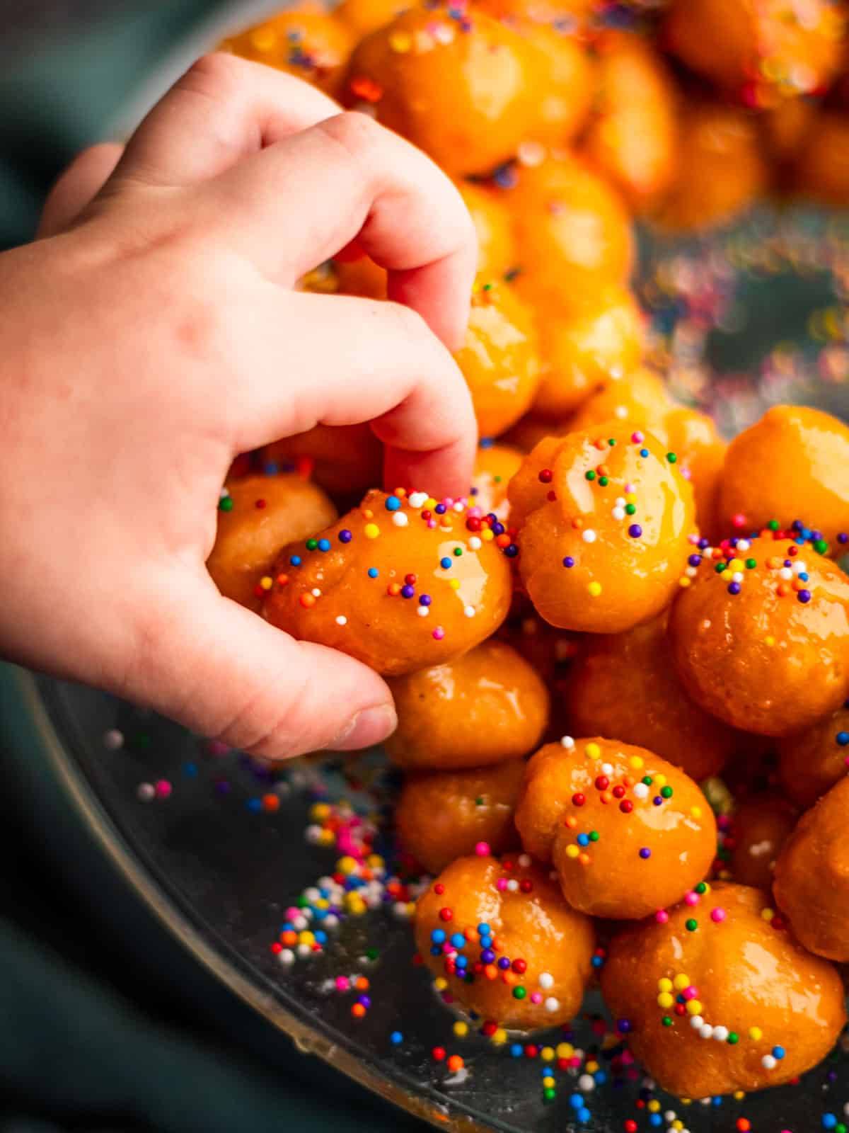 hand grabbing a struffoli ball covered in sprinkles off of a platter.