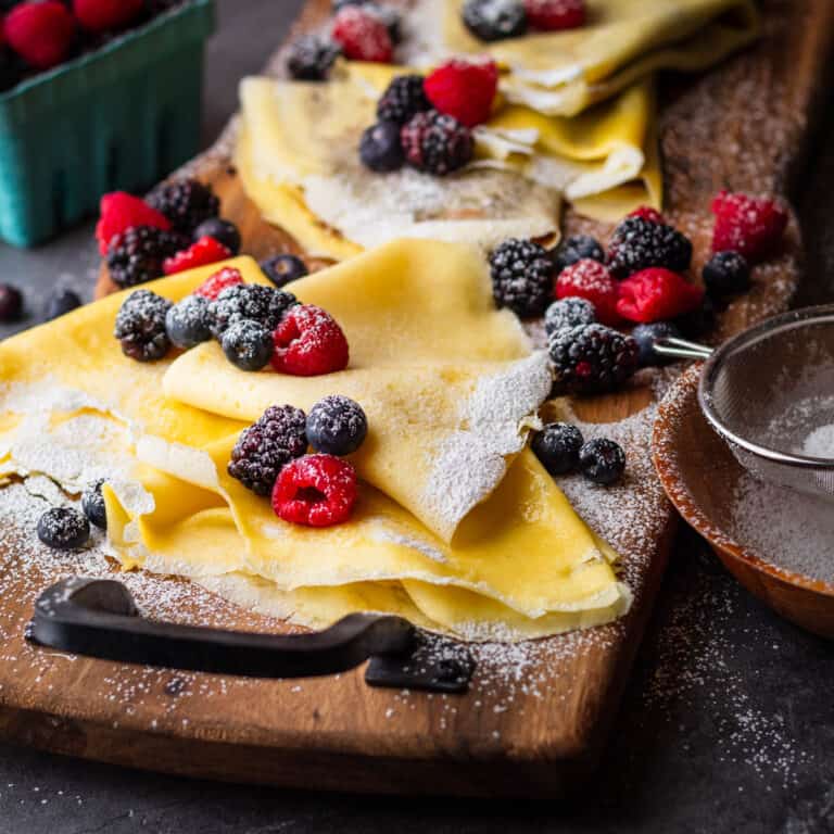 crepes folded on a wooden tray with berries and powdered sugar sprinkled on them.