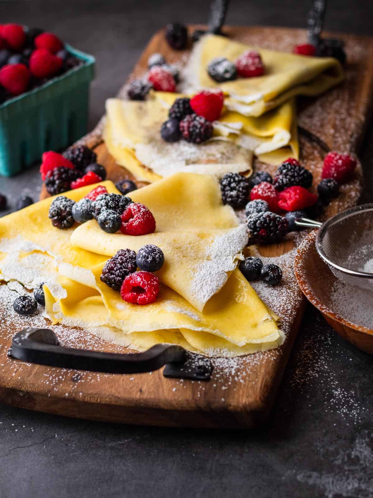 folded crepes on a wooden tray topped with fresh berries and powdered sugar on and around.
