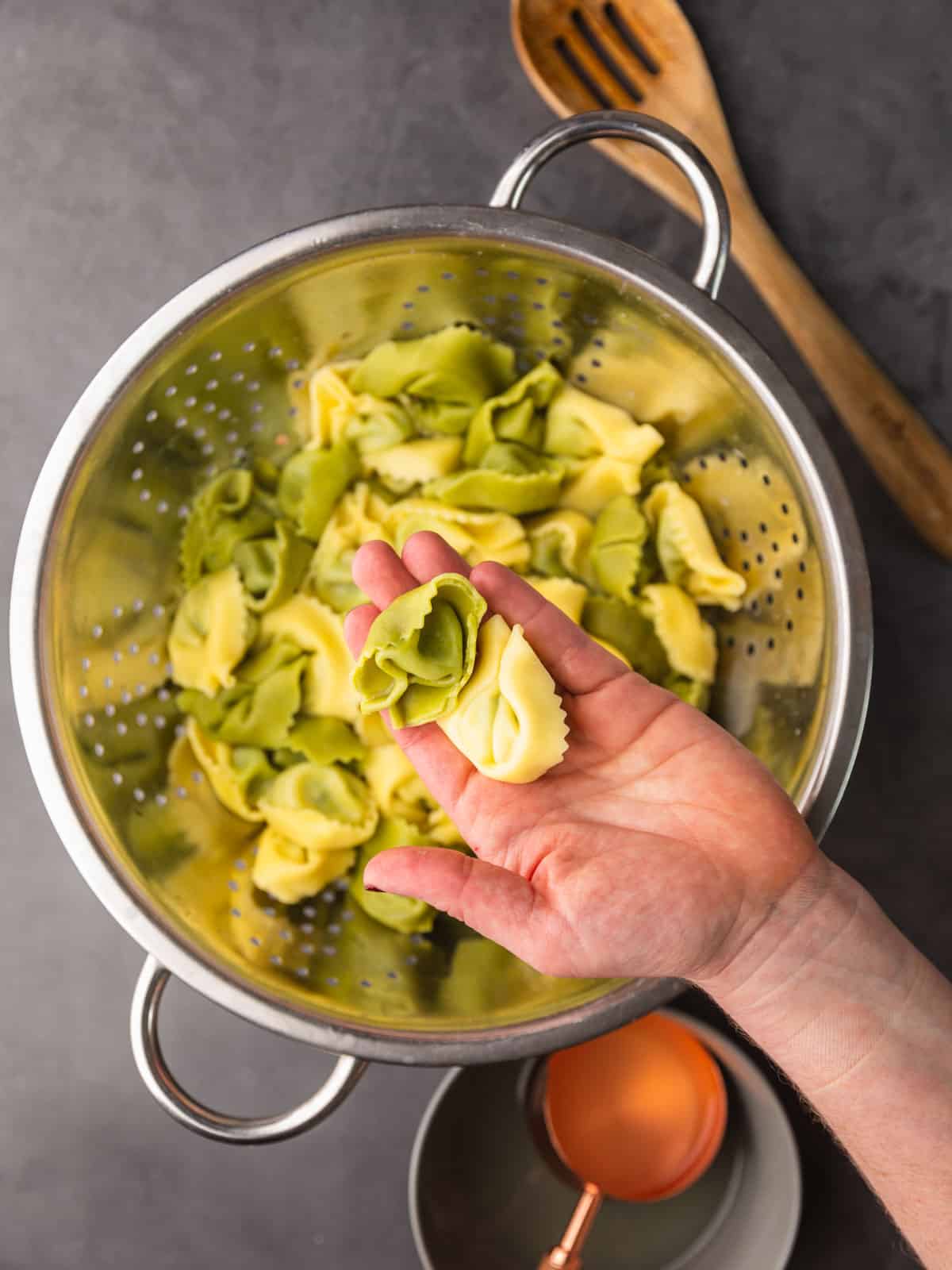 hand holding cooked pieces of green and white tortellini pasta over a colander.