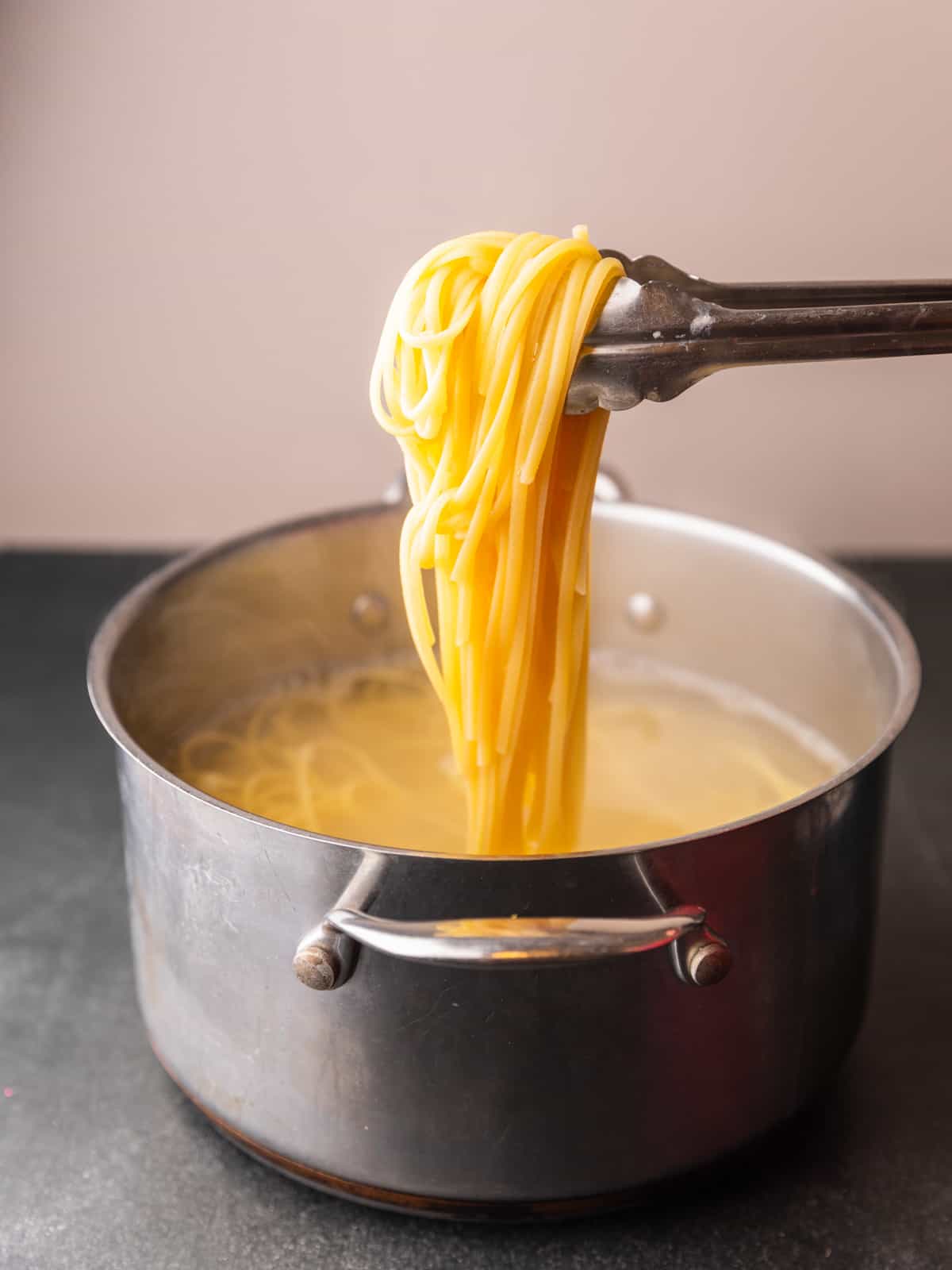 tongs holding a bunch of cooked linguine pasta over a pot of pasta cooking in water.