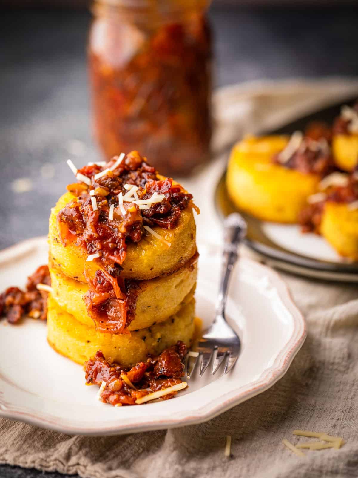 stack of 3 fried polenta cakes on a plate with tomato bacon jam and parmesan.