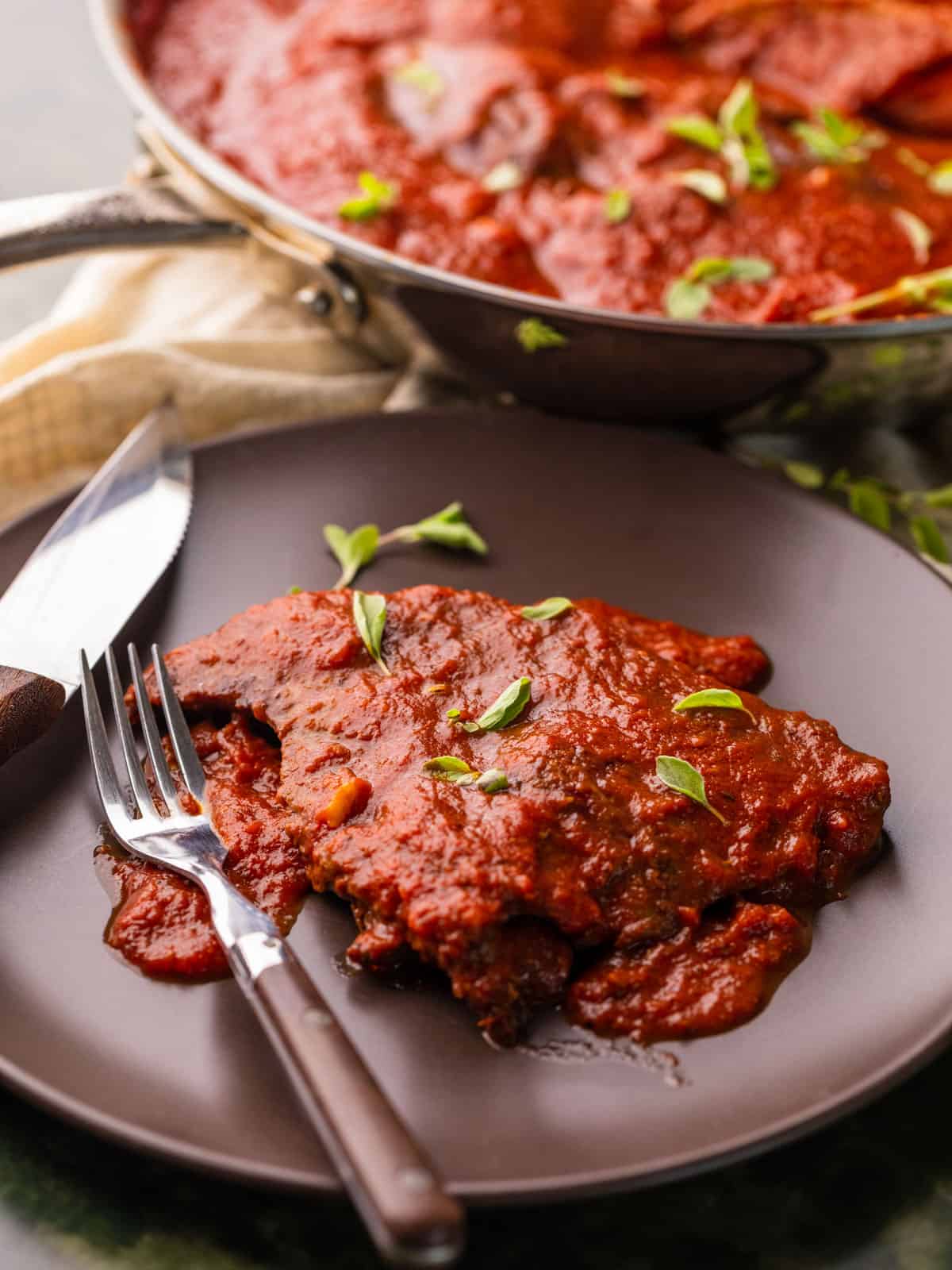 steak in red sauce on a plate with a fork and steak knife and fresh oregano.