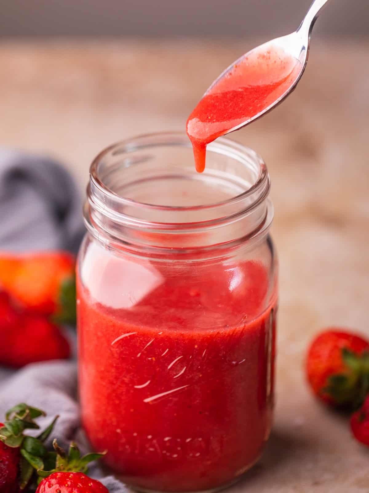 strawberry puree dripping off a spoon into a mason jar of puree with fresh strawberries around.