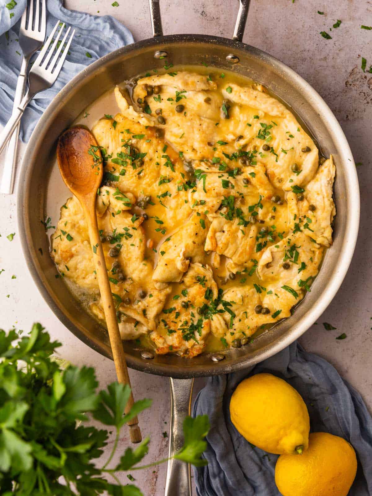 skillet with chicken piccata with capers in it with a wooden spoon and lemons around.