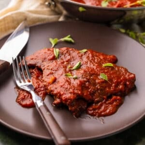steak covered in red sauce on brown plate with fresh oregano, a fork and steak knife.