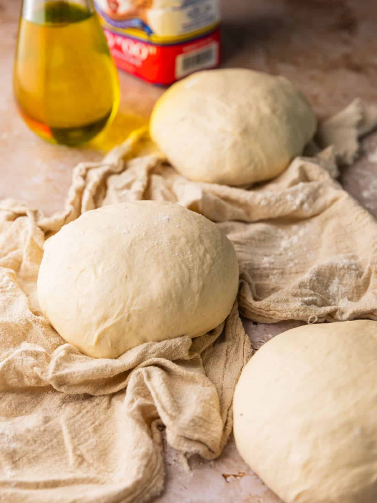 balls of pizza dough on a table with a towel with oil and flour behind them.
