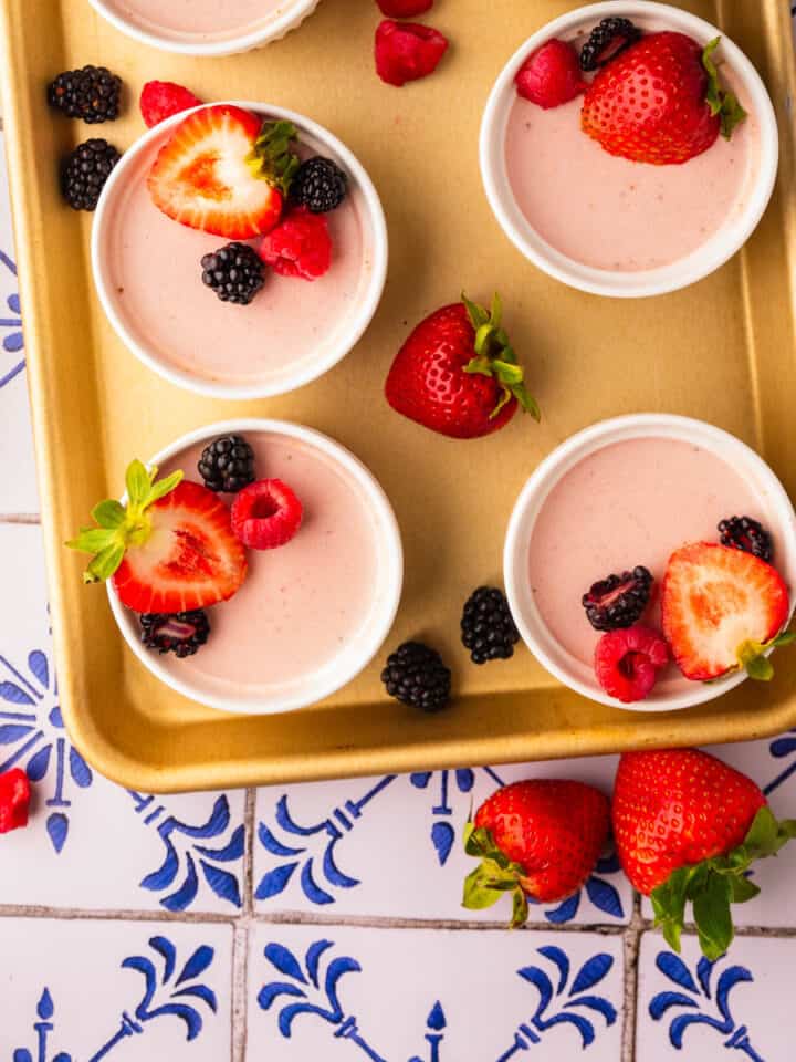 ramekins of strawberry panna cotta topped with fresh berries on a gold sheet pan.