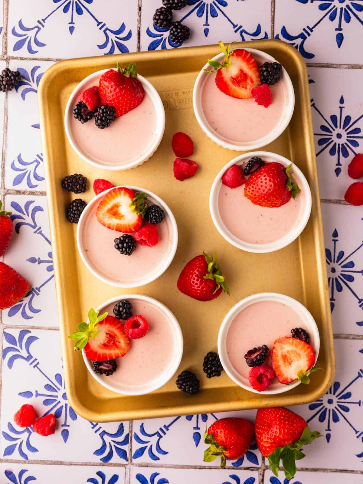 strawberry panna cotta in white ramekins topped with berries on a gold sheet pan.