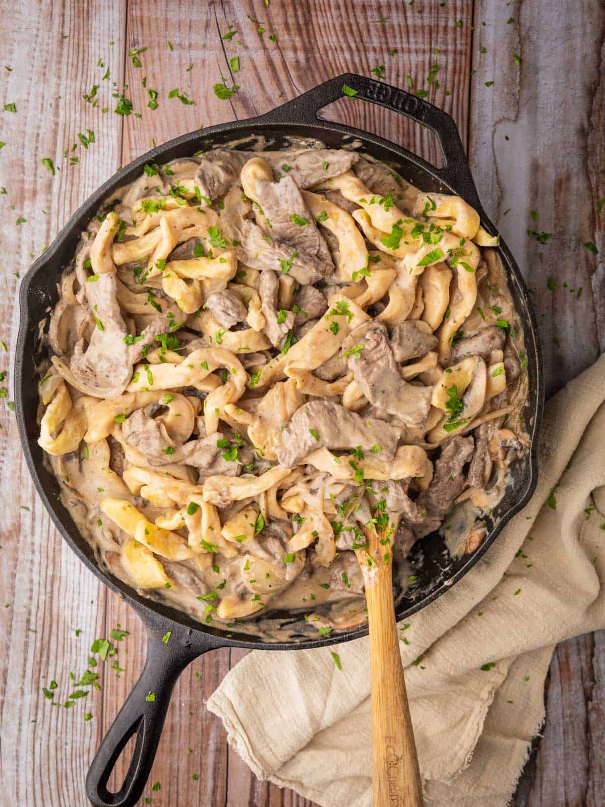 cast iron skillet of beef stroganoff with egg noodles with a wooden spoon in it.