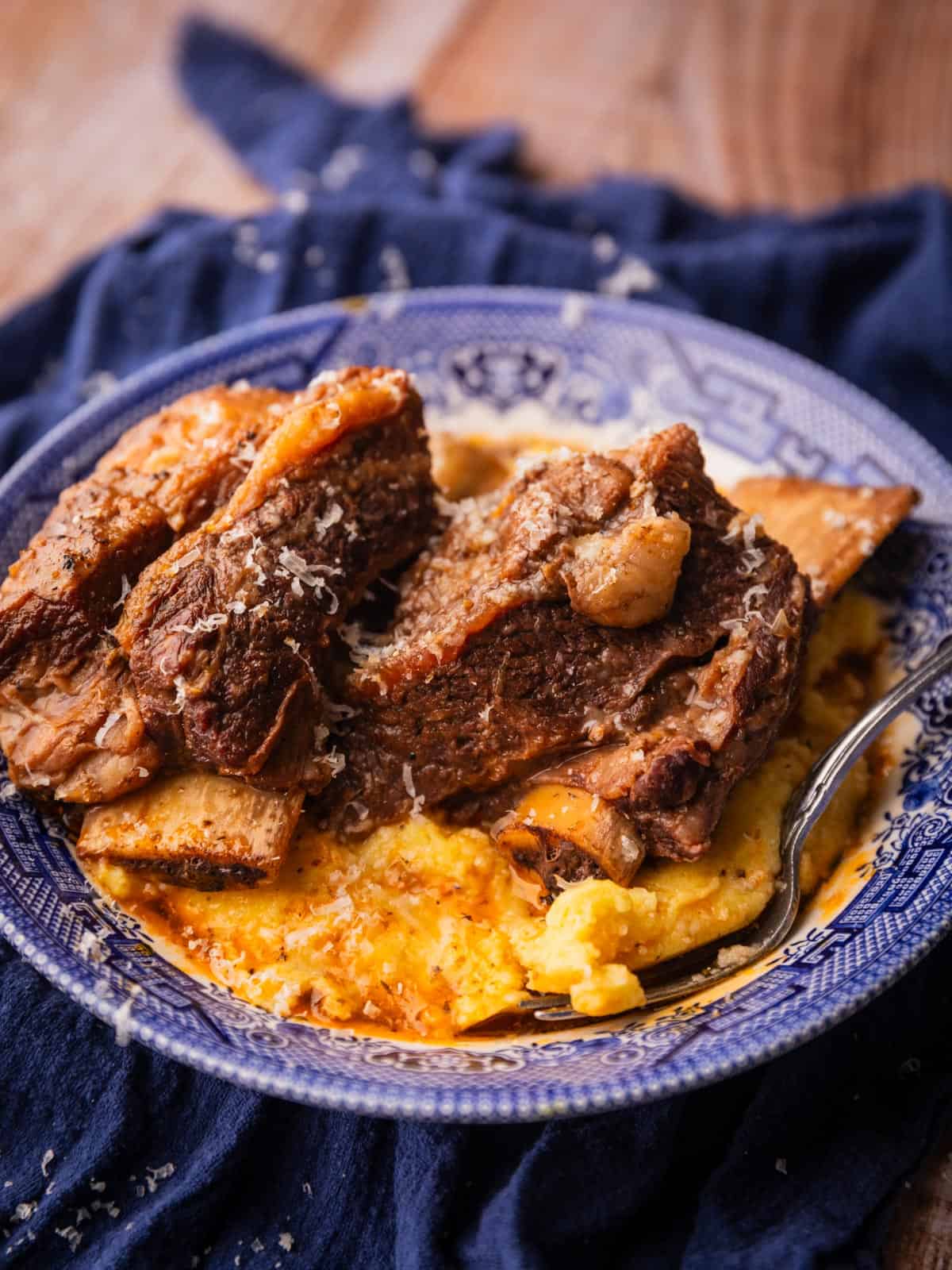 cooked beef short ribs in a bowl over polenta with a fork in it.