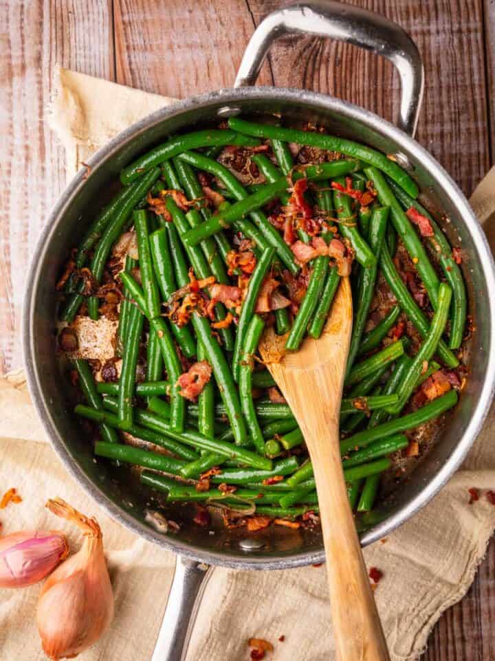 green beans with bacon and shallots in a silver skillet with a wooden spoon.
