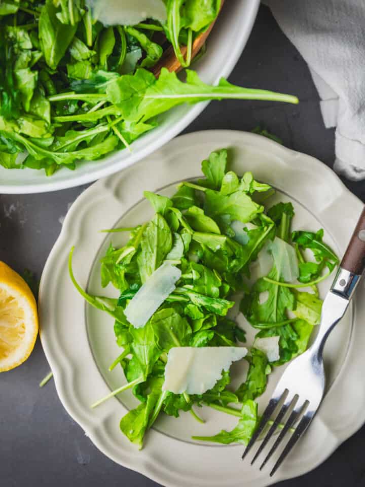 arugula salad with parmesan on plate and in bowl with cut lemon next to it.