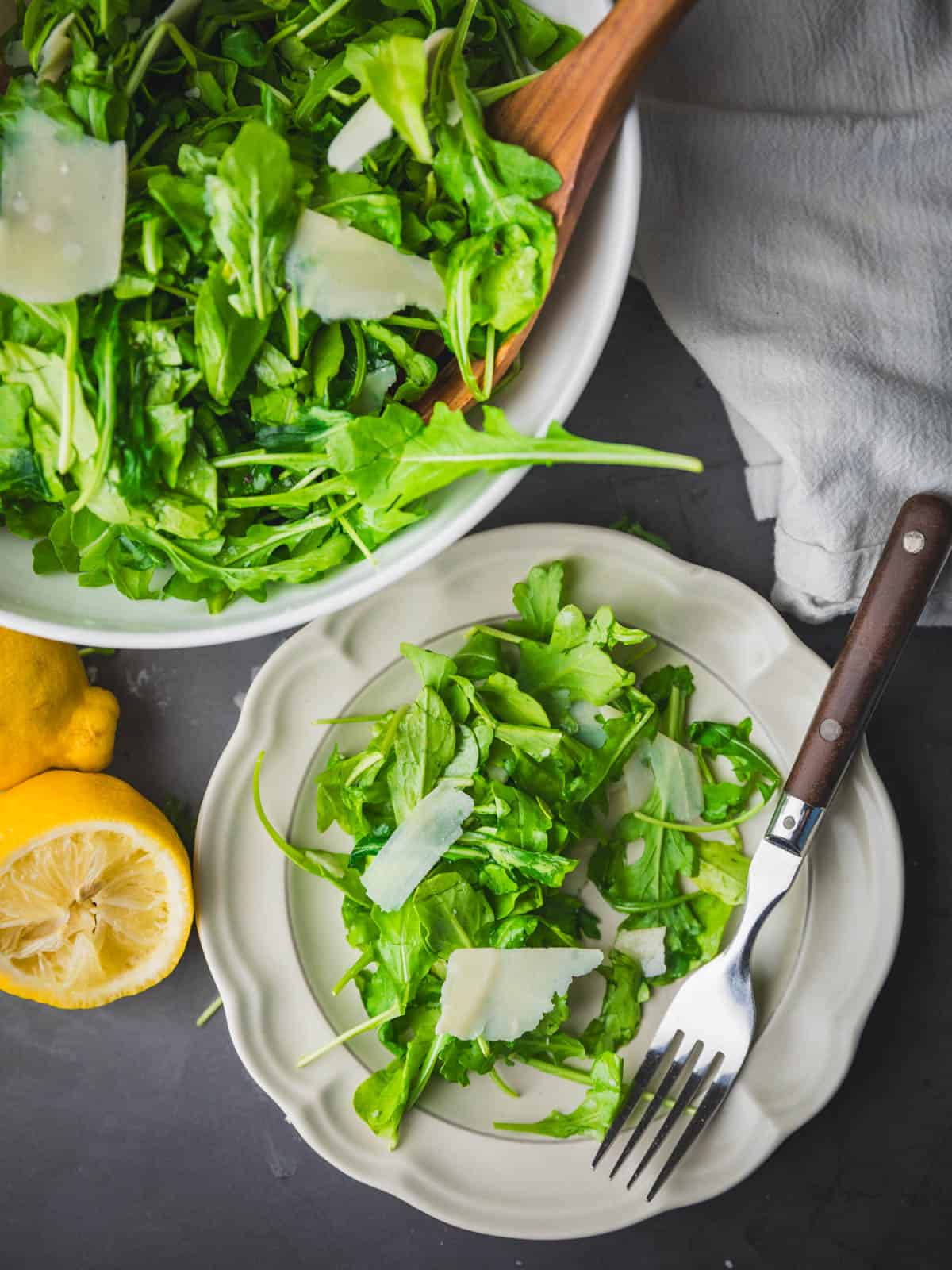 arugula parmesan salad in bowl and on plate with fork next to cut lemon.