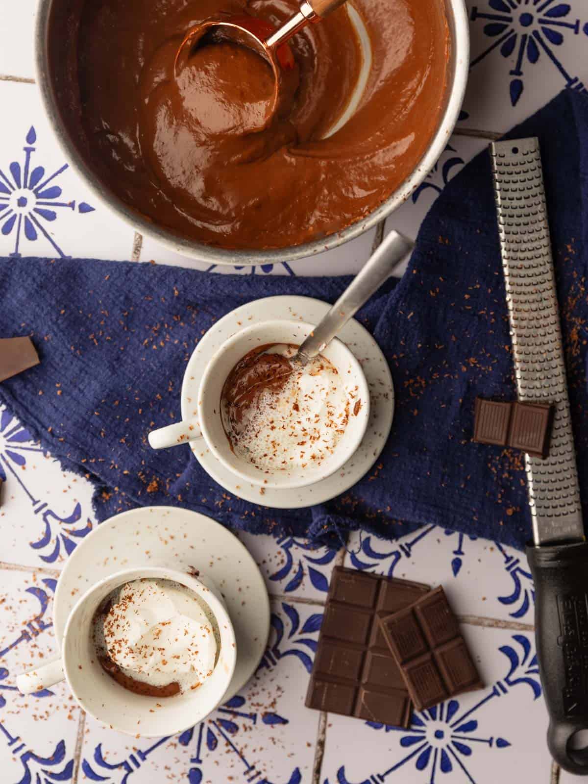 pot of italian hot chocolate next to two cups of it with cream and chocolate.