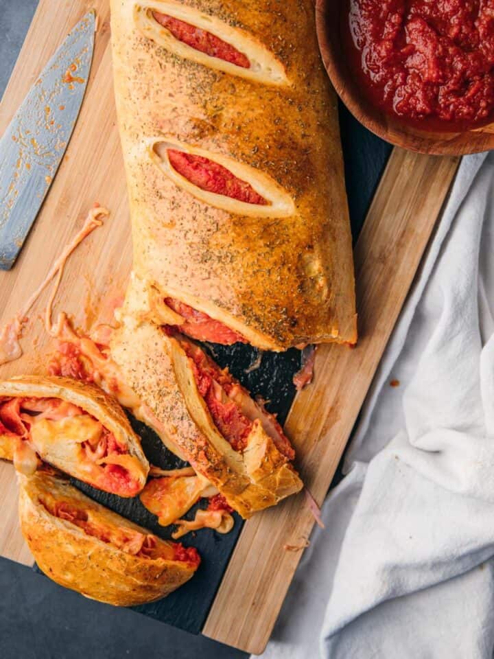 stromboli partially sliced on a cutting board next to a knife and bowl of pizza sauce.