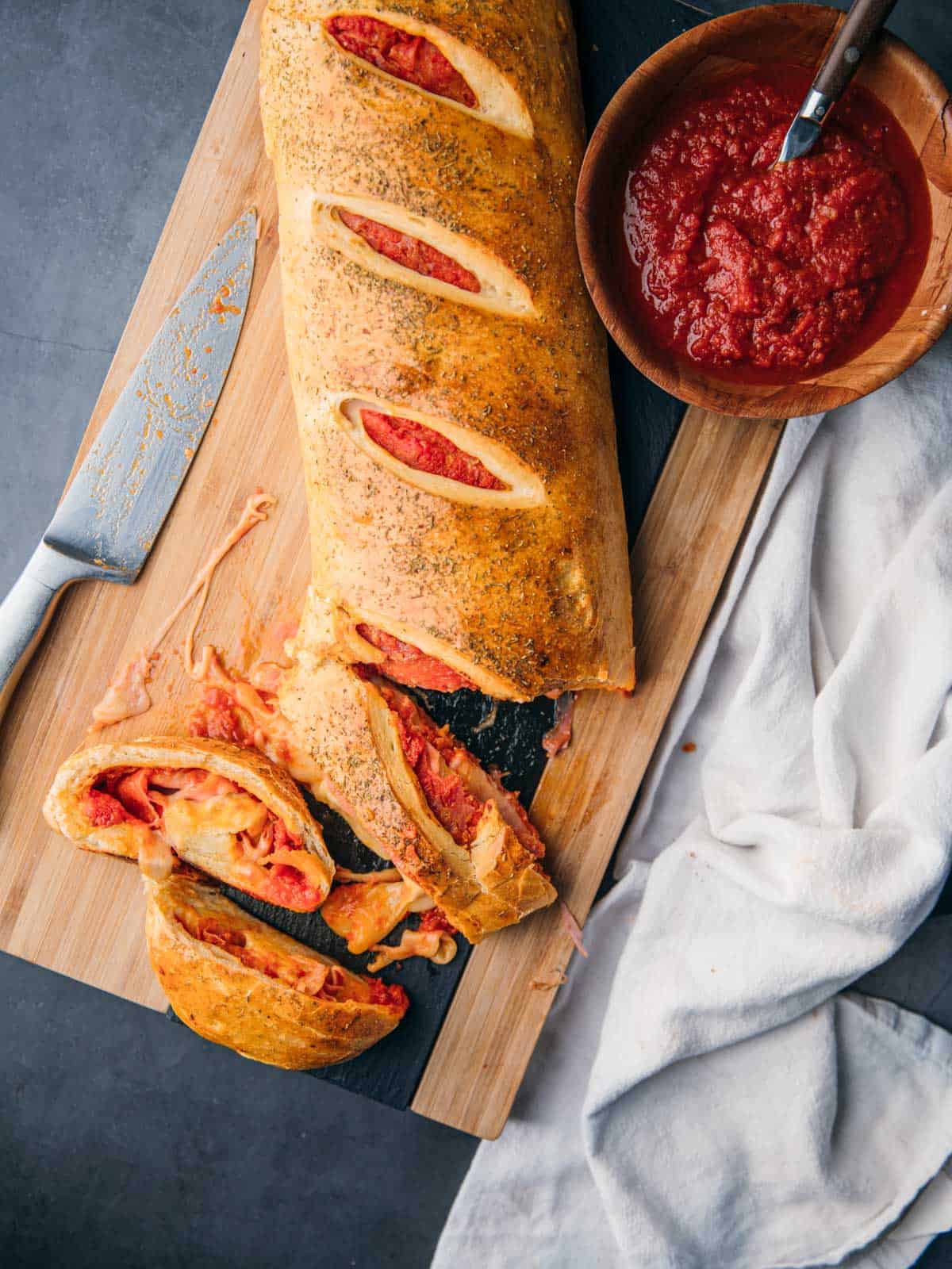 stromboli sliced on a cutting board next to a knife and bowl of pizza sauce.