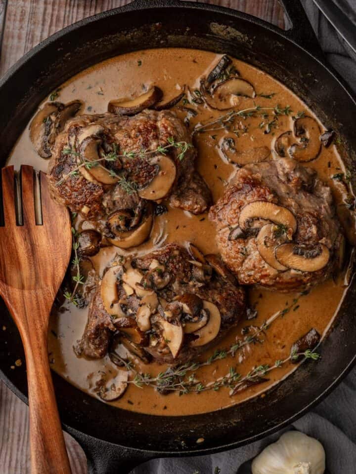 steaks in a mushrooms sauce in a cast iron skillet with fresh thyme sprigs on top.