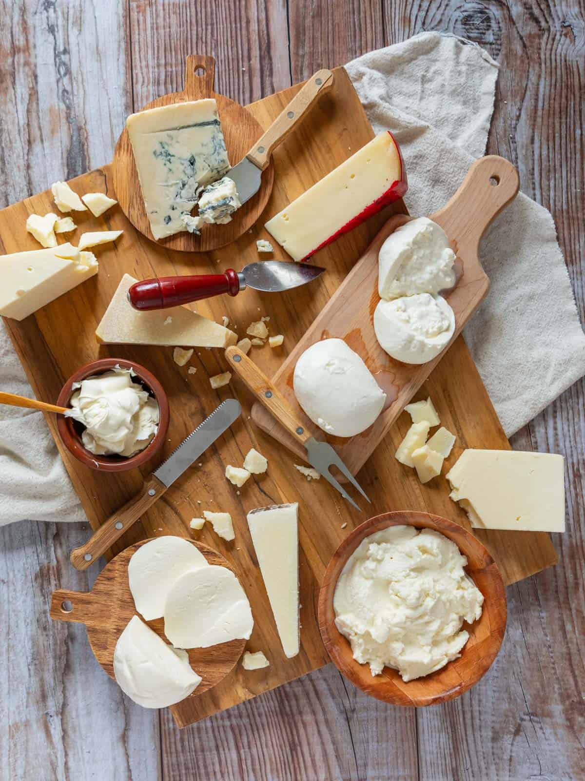 hard and soft italian cheeses in wedges and bowls on a cutting board with cheese knives and forks.
