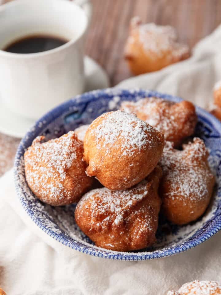 zeppole donuts sprinkled with powdered sugar in a blue and white bowl next to a cup of coffee.