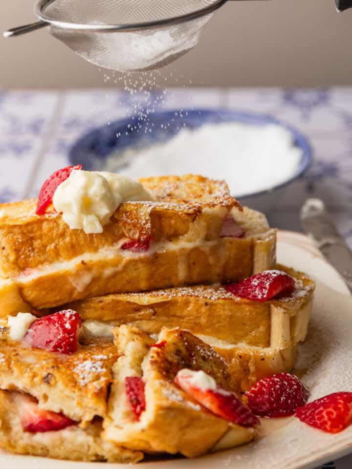 powdered sugar being sprinkled over a stack of stuffed french toast with fresh strawberries.