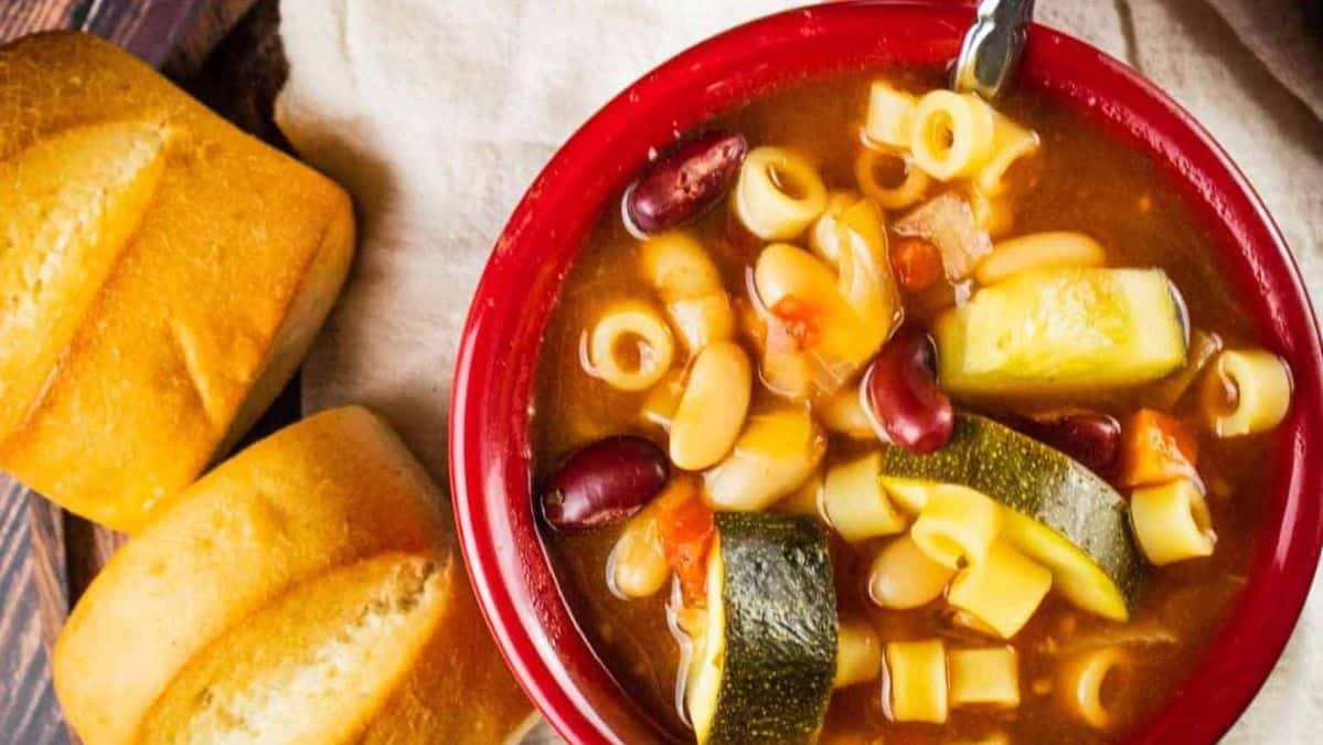 bowl of minestrone soup with a spoon and rolls sitting next to it.