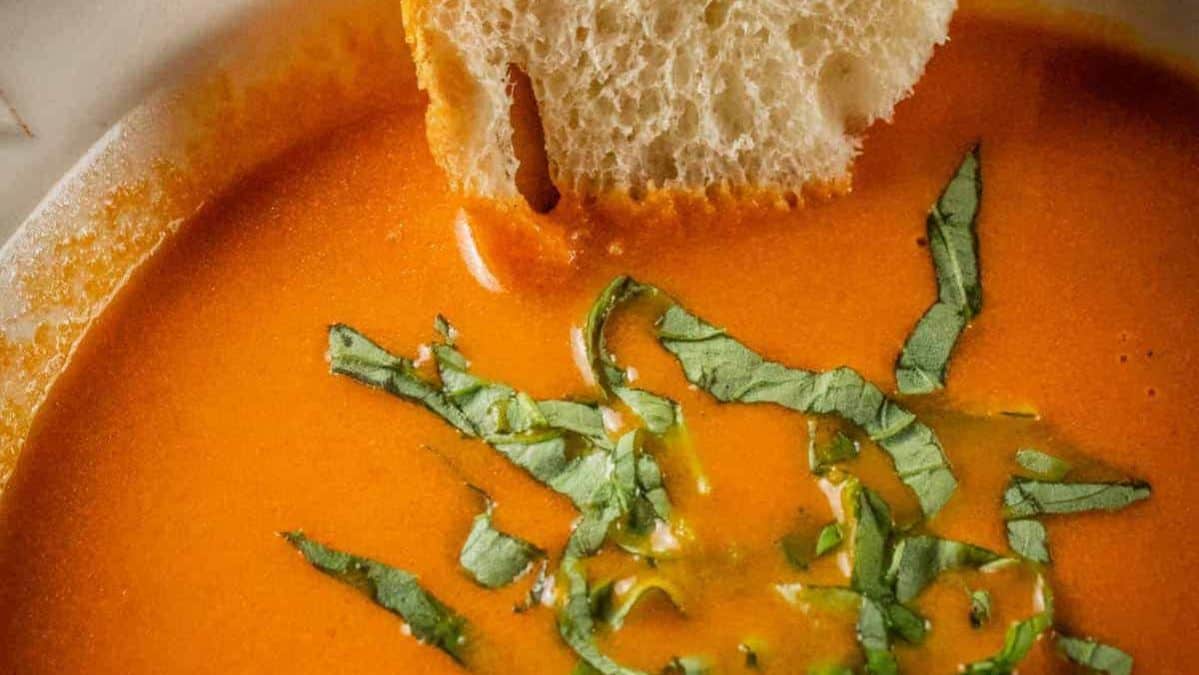 bowl of creamy tomato soup topped with sliced fresh basil and piece of bread sitting in it.