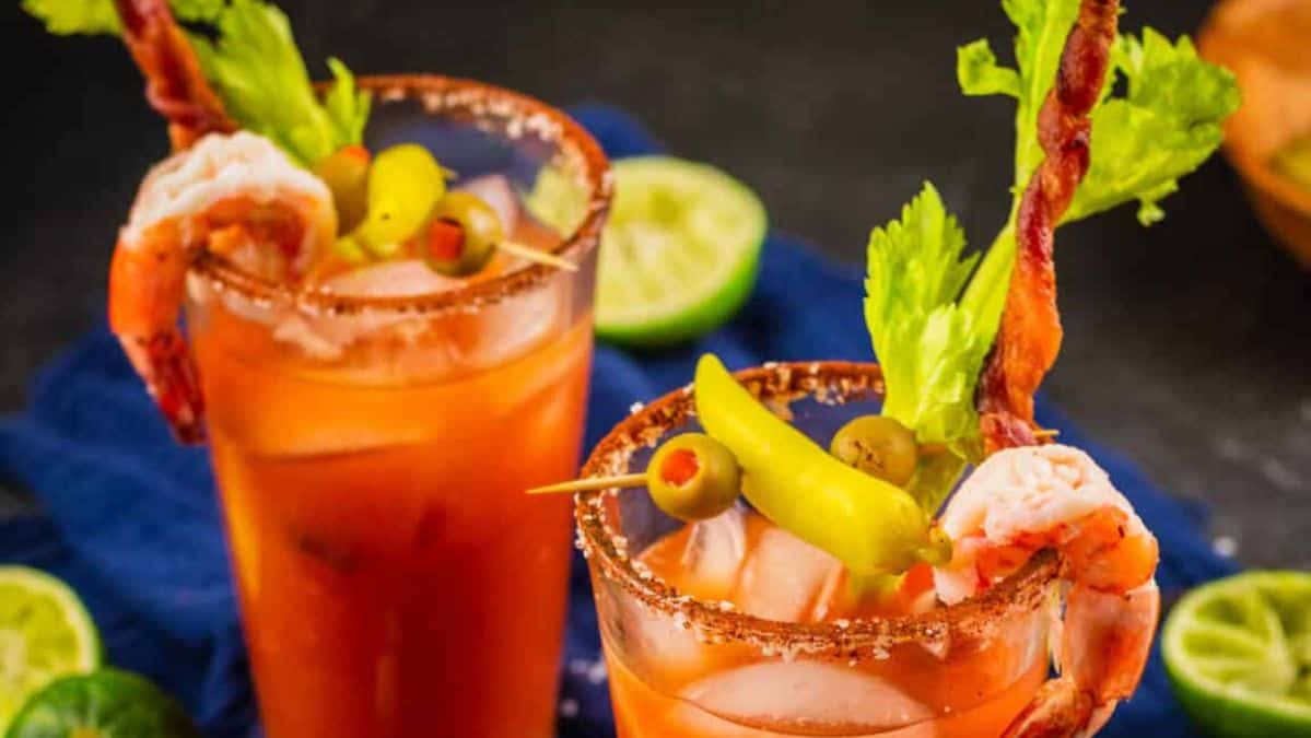 two bloody marys in clear glasses with a spiced rim garnished with twisted bacon, shrimp, peppers, olives and celery.