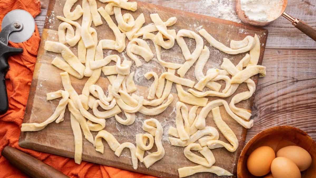 Uncooked egg noodles on a cutting board next to whole eggs and a cup of flour.