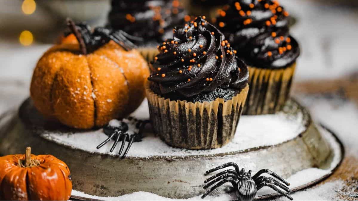 Chocolate cupcakes with black frosting and orange sprinkles on a platter next to fake spiders and fake pumpkins.