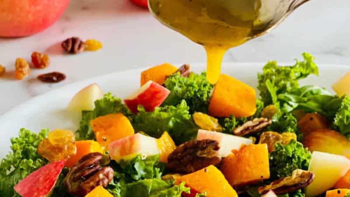 Yellow dressing, dripping off of a spoon onto a salad with apples, butternut squash, and kale.