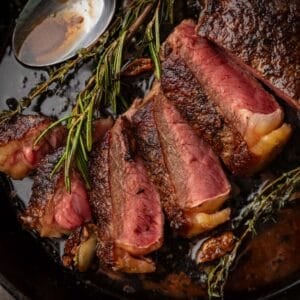 Shot of a medium rare cooked steak sliced in a cast-iron skillet next to sprigs of fresh herbs and a spoon with butter sauce in it.