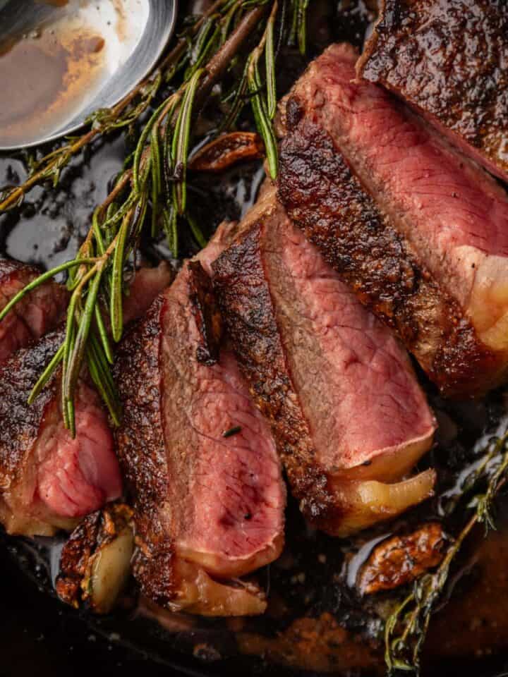 Shot of a medium rare cooked steak sliced in a cast-iron skillet next to sprigs of fresh herbs and a spoon with butter sauce in it.