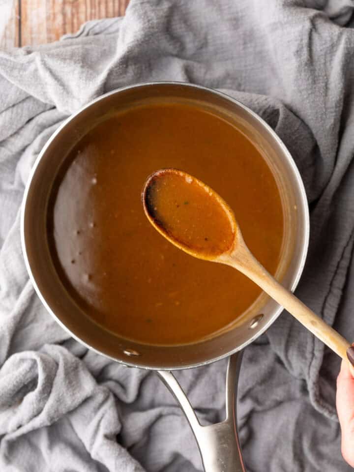 hand holding a wooden spoon filled with brown gravy over a pot of gravy.