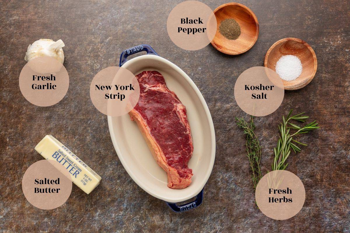 Head of garlic, a NY strip steak in a dish, a stick of butter, wooden dishes of black pepper and kosher salt, with fresh herb sprigs lying on a table.