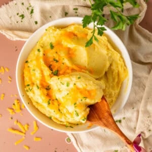 Wooden spoon holding a scoop of mashed potatoes with melted cheese and fresh herbs on top.