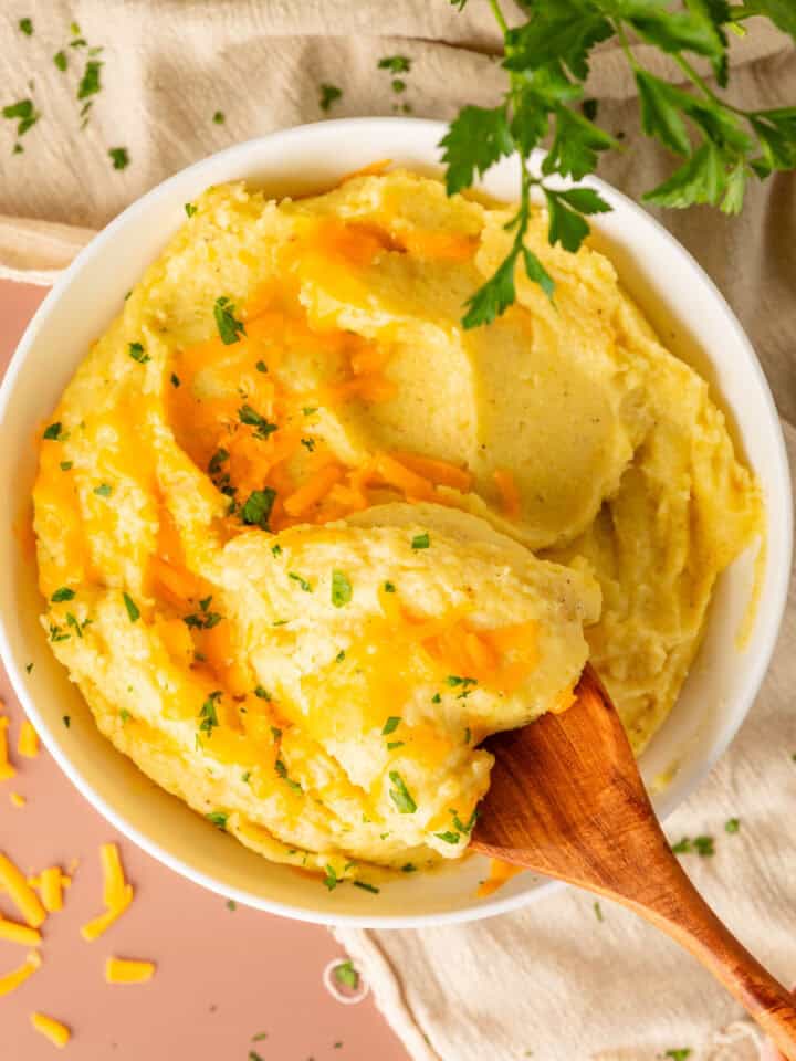 Wooden spoon holding a scoop of mashed potatoes with melted cheese and fresh herbs on top.