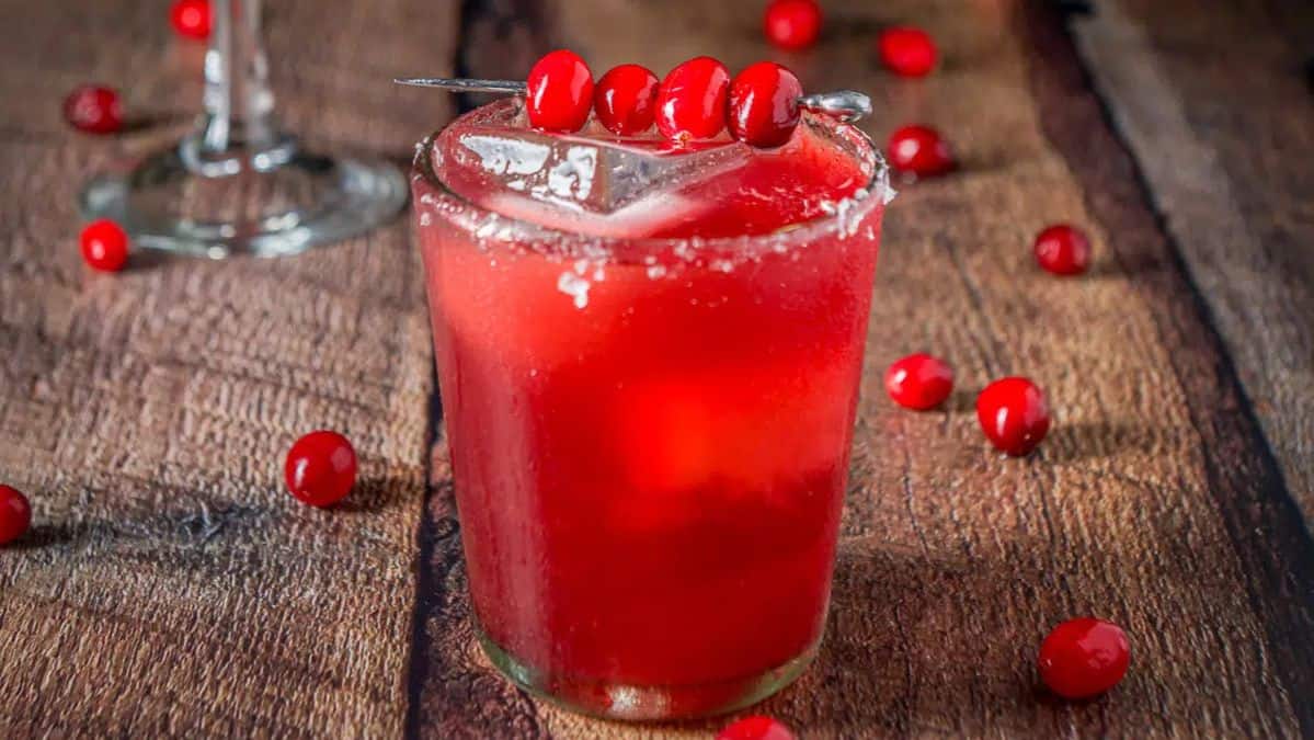 Clear glass with a red colored drink it with a salted rim garnished with whole cranberries.