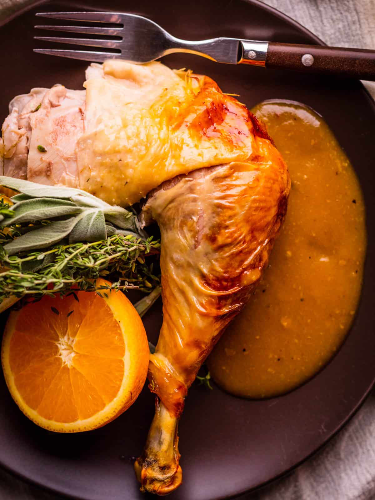 Turkey thigh and drumstick on a plate with with gravy, a fork, fresh herbs, and an orange half.