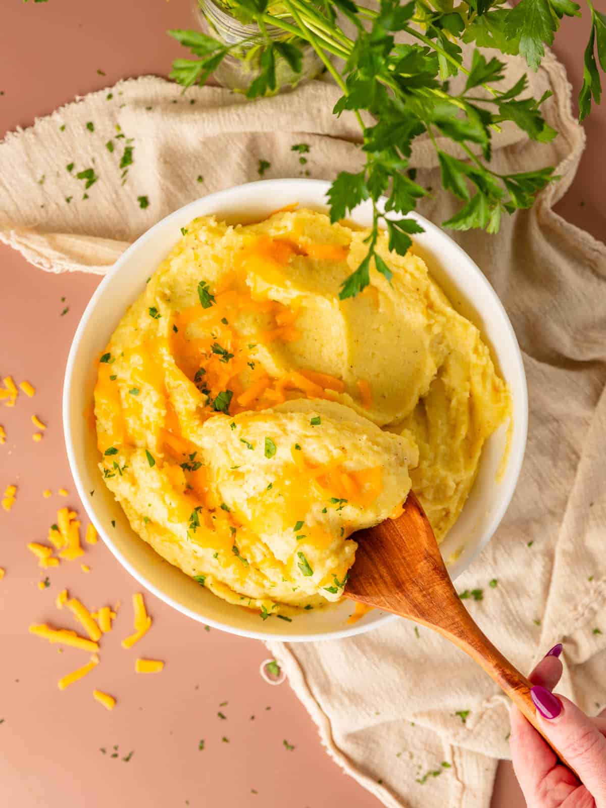 A bowl of mashed potatoes with melted cheese and herbs on top with a hand scooping some of them with a wooden spoon.