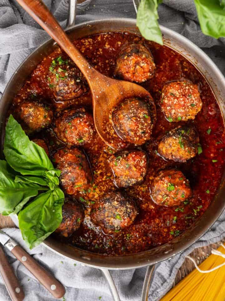 Meatballs in marinara in a skillet with a wooden spoon, garnished with fresh herbs and Parmesan cheese.