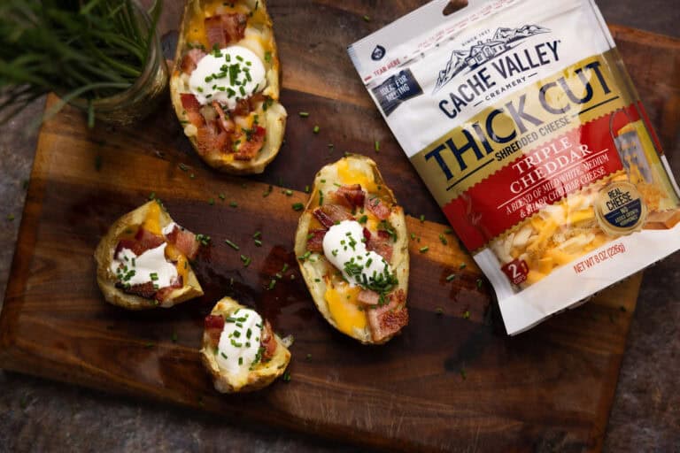Three loaded potato skins, one split in half next to a bag of shredded Cache Valley cheese.