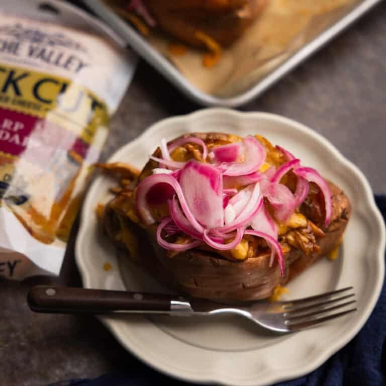 A BBQ chicken stuffed sweet potato topped with cheese and pickled red onions on a plate with a fork.