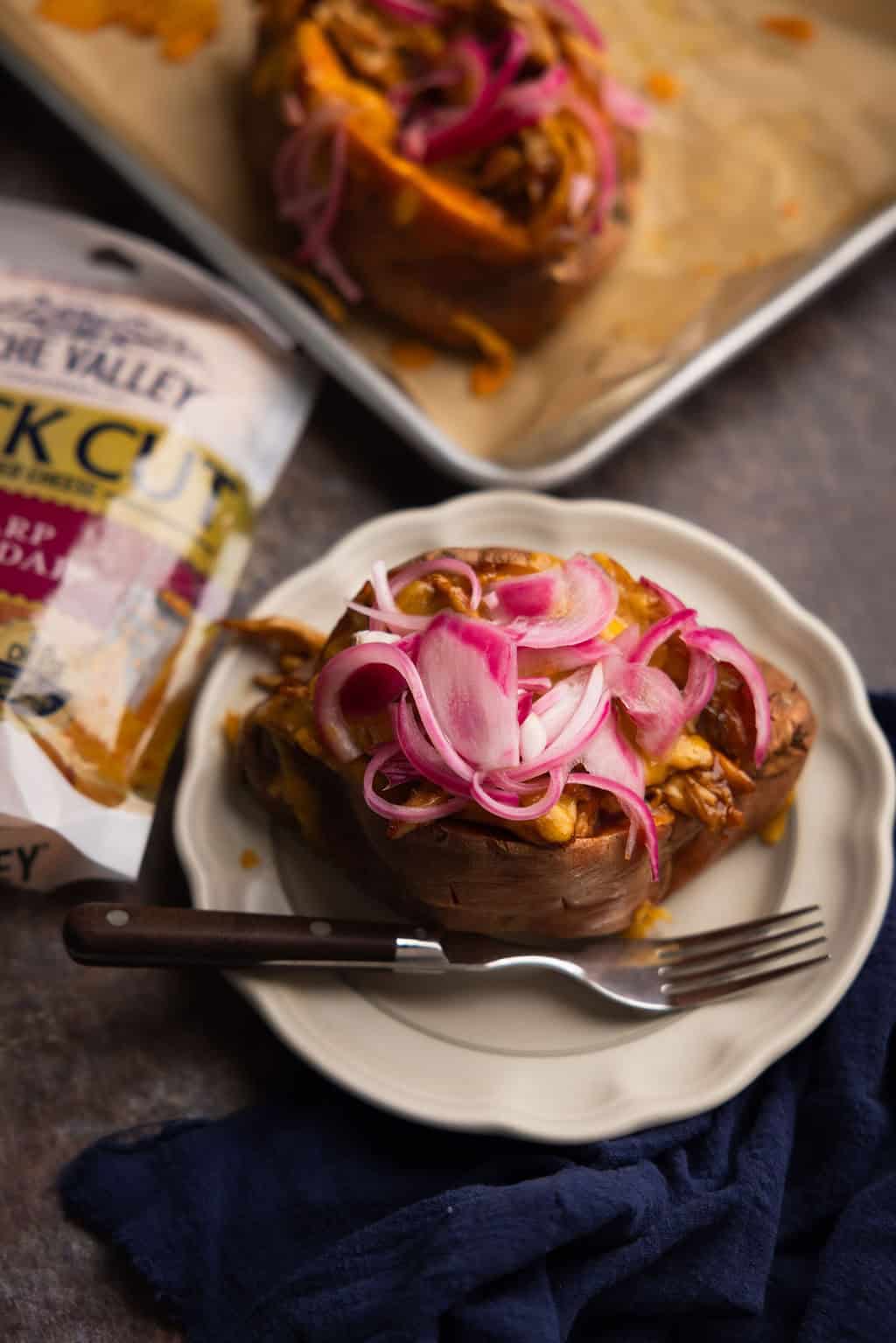 A BBQ chicken stuffed sweet potato topped with cheese and pickled red onions on a plate with a fork. 